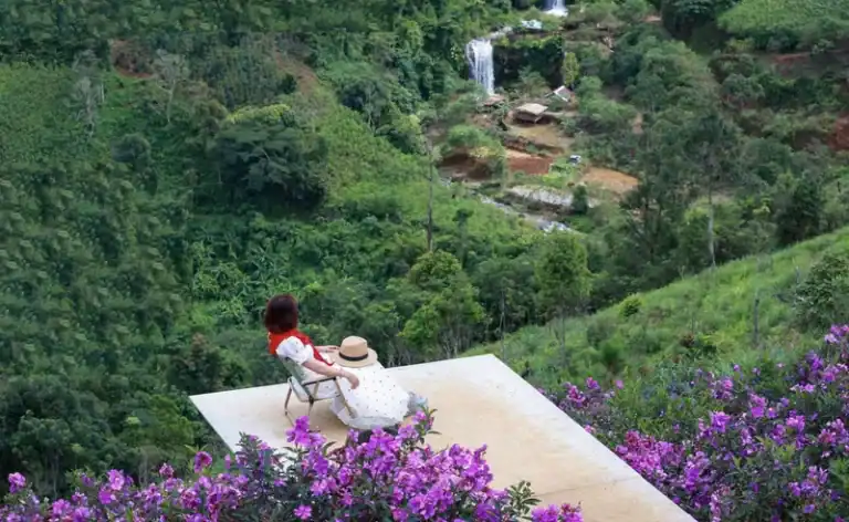 Phụ nữ mặc váy trắng ngồi trên ghế sân hiên nhìn ra thung lũng xanh, xung quanh là hoa mua Măng Đen nở tím, thác nước xa xa