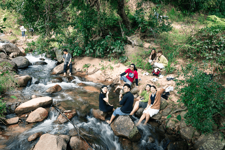 đoàn khách ngồi trải nghiệm bắt cá suối và tắm suối bên bờ suối có nhiều đá xung quanh là rừng nguyên sinh tại Kon Tu Ma Măng Đen trong tour trekking Kon Tu Ma