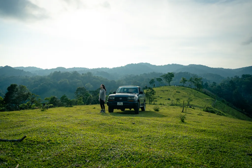 một cô khách du lịch trong chuyến đi tham quan tại đồi bò Măng Cành trong một chuyến tham quan Măng Đen cùng xe 2 cầu của Măng Đen Discovery với rừng cây và đồi núi trùng điệp sau lưngnh bên đồi cỏ, ánh sáng rất đẹp