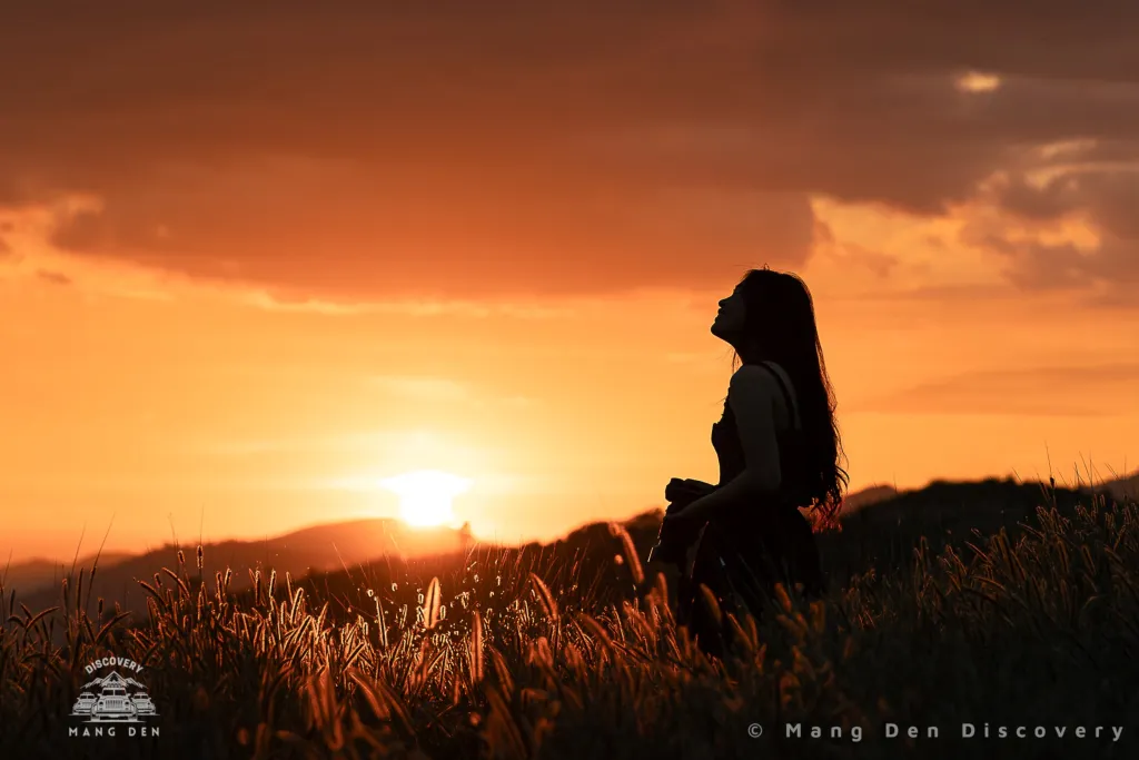 cô gái cầm máy ảnh tại đồi cỏ măng đen với mặt trời đang lặn sau đồi núi phía xa lúc hoàng hôn